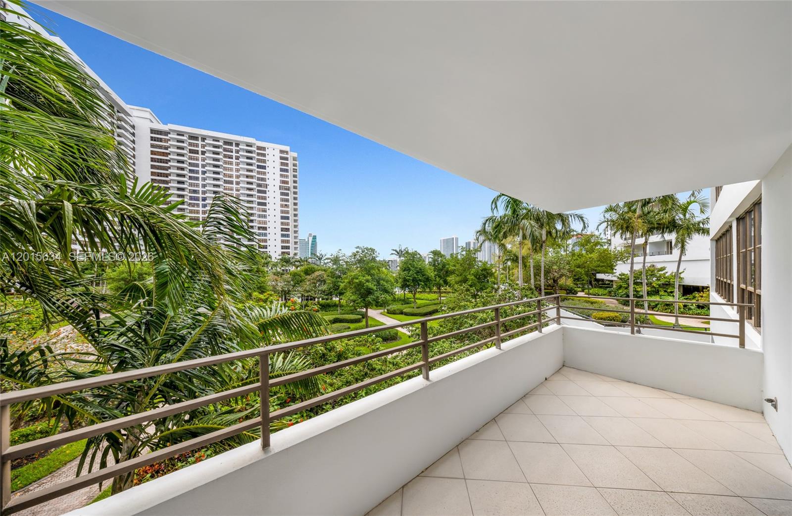 A balcony with a tiled floor and metal railing overlooks a landscaped garden and buildings — 600 Three Islands Blvd # 212, Hallandale Beach FL 33009, HALLANDALE BEACH, FL