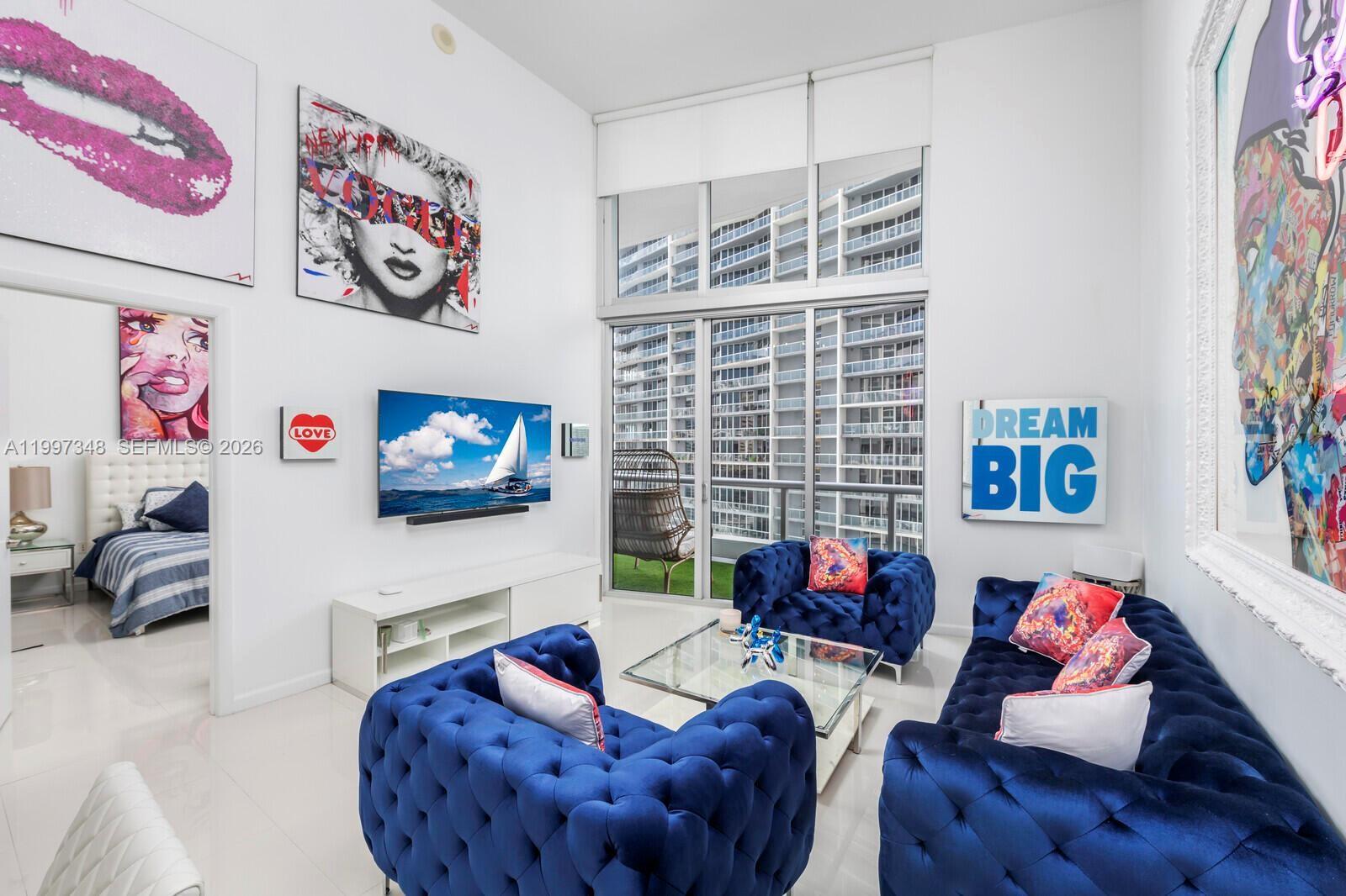 A modern living room features blue tufted furniture, pop art, and floor-to-ceiling windows — 475 Brickell Ave # 2811, Miami FL 33131, MIAMI, FL