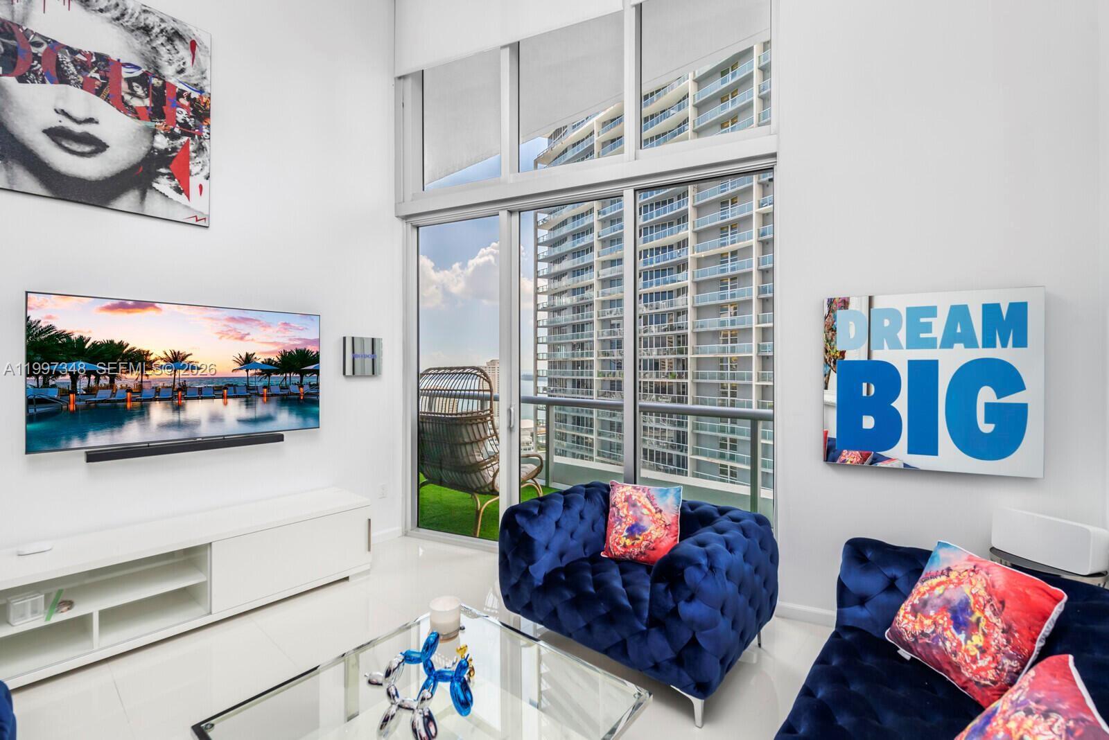 A modern living room features blue tufted furniture, a wall-mounted television, and floor-to-ceiling windows — 475 Brickell Ave # 2811, Miami FL 33131, MIAMI, FL