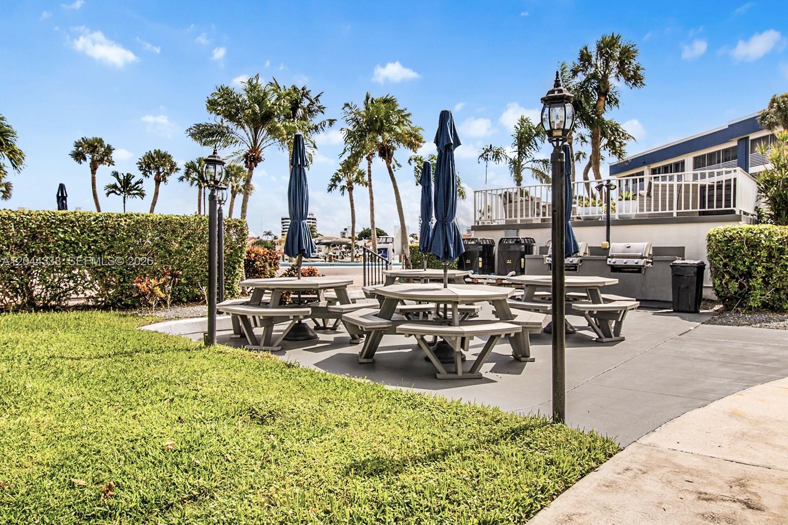 An outdoor patio area features several picnic tables with blue umbrellas and a grill station — 3401 Spanish Trl # 454, Delray Beach FL 33483, DELRAY BEACH, FL
