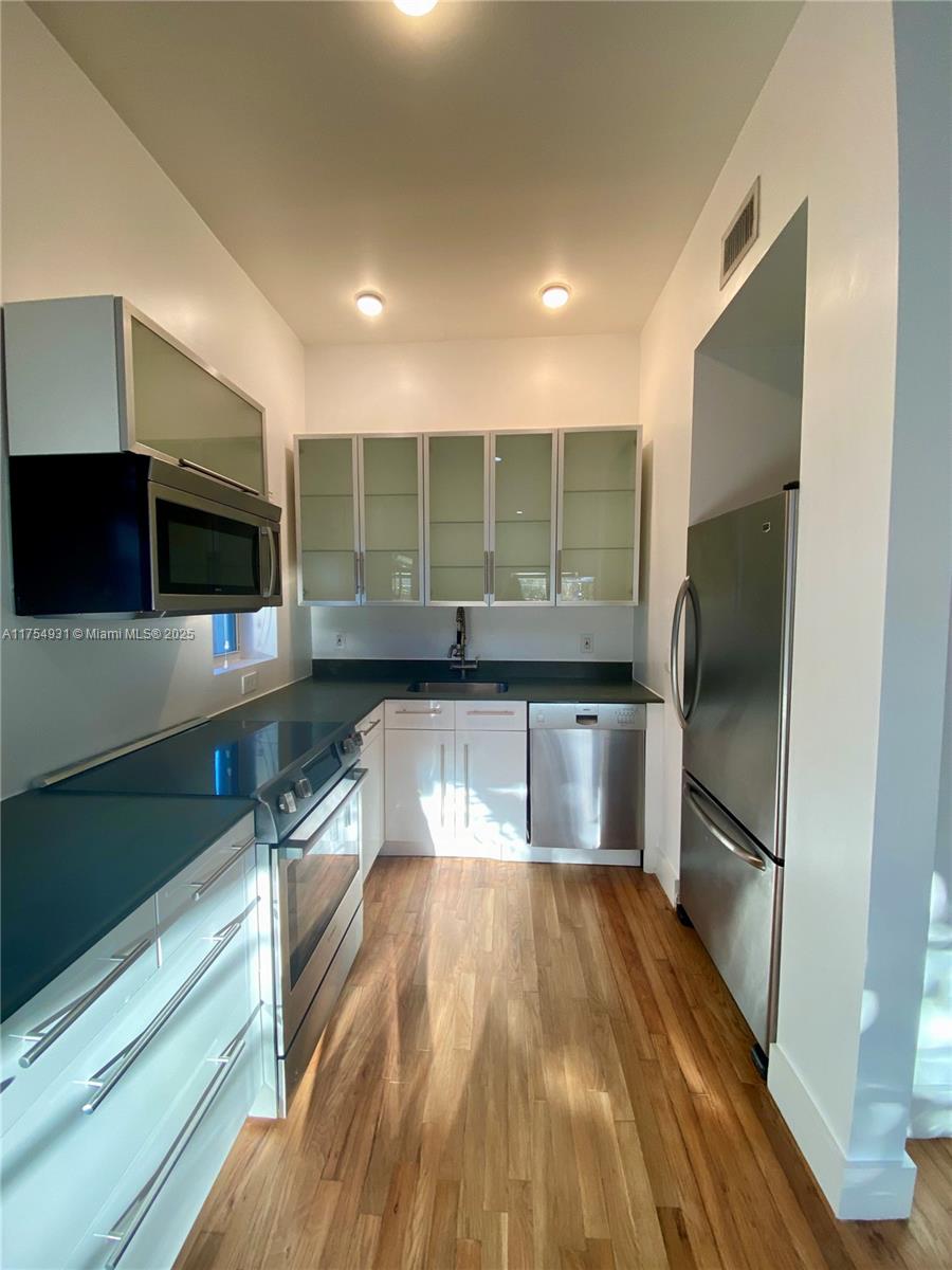 A galley kitchen features white cabinets, stainless steel appliances, dark countertops, and wood flooring — 929 Michigan Ave # 5, Miami Beach FL 33139, MIAMI BEACH, FL