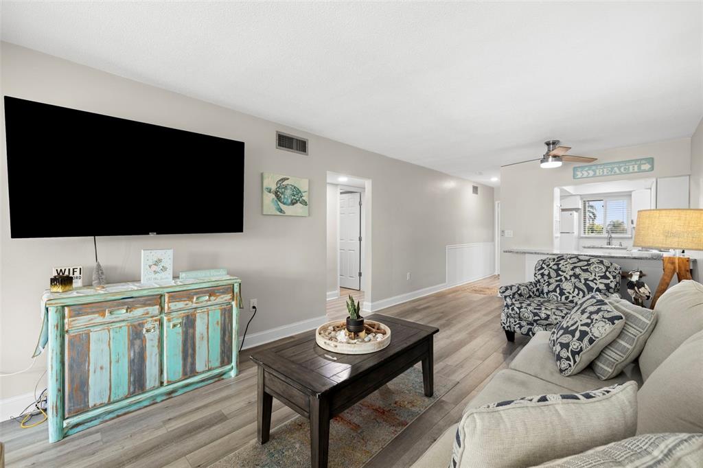 A living room features a large television, a rustic turquoise cabinet, and wood-look flooring — 6767 SUNSET WAY UNIT 206, ST PETE BEACH, FL