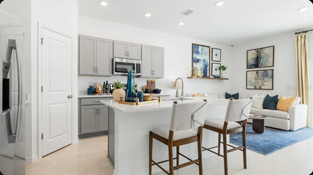 A kitchen with gray cabinets and a white island features bar stools and an adjacent living area — 7187 GRAND FOREST WAY, NOKOMIS, FL