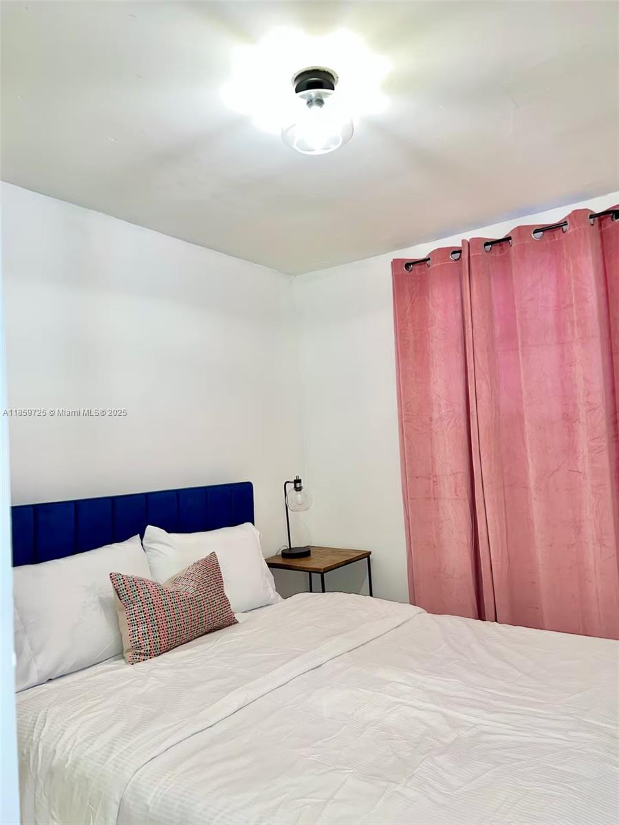 A bedroom features a blue headboard, white bedding, a small side table, and pink curtains — 2701 NW 24th Ct # R, Miami FL 33142, MIAMI, FL