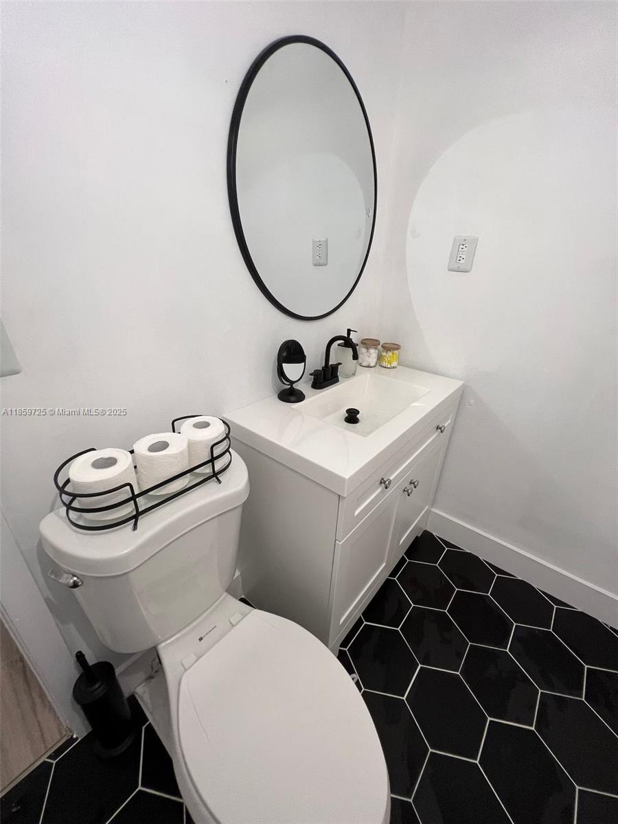 A small bathroom features a white vanity, oval mirror, toilet, and black hexagonal floor tiles — 2701 NW 24th Ct # R, Miami FL 33142, MIAMI, FL