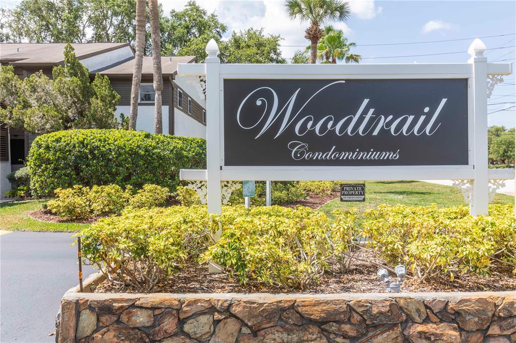 A white sign for Woodtrail Condominiums stands in front of a building and landscaping — 4008 PASSPORT LANE UNIT 104, NEW PORT RICHEY, FL