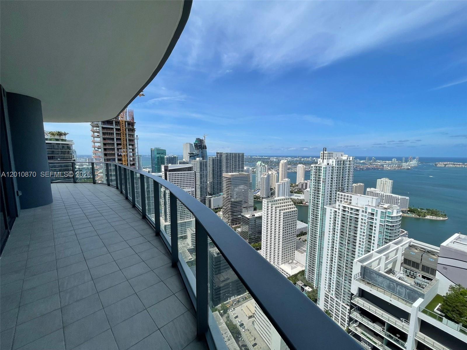 A high-rise balcony with gray tile flooring overlooks a dense city skyline and the ocean — 1000 Brickell Plaza # 5908, Miami FL 33131, MIAMI, FL