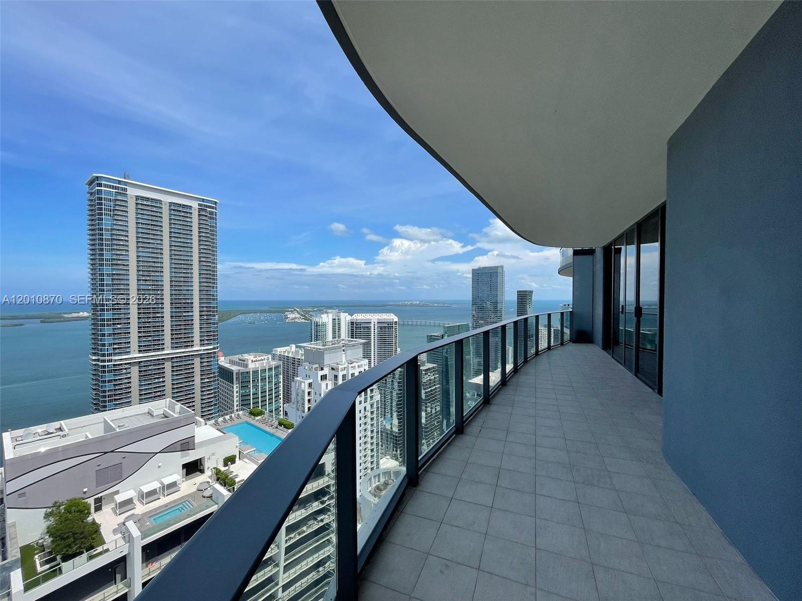 A curved balcony with glass railings overlooks a city skyline and a large body of water — 1000 Brickell Plaza # 5908, Miami FL 33131, MIAMI, FL