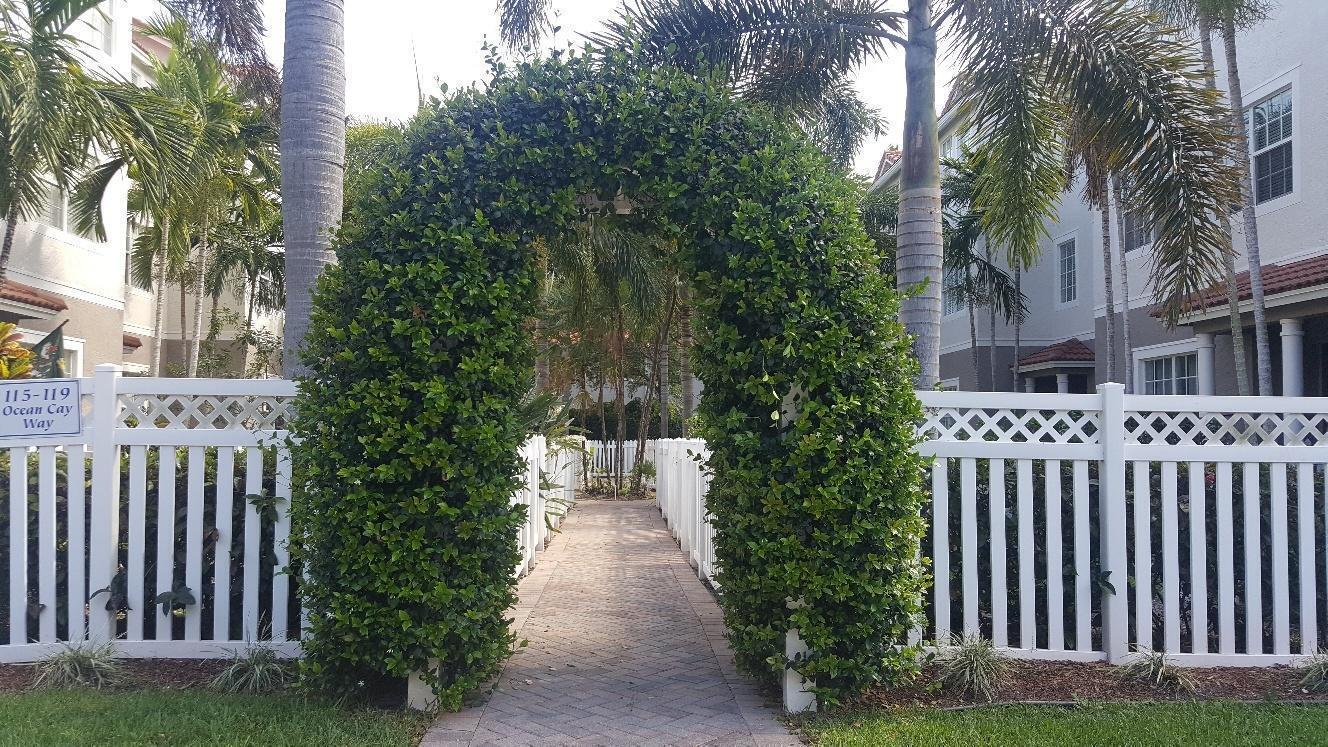 A brick walkway leads through a leafy green archway framed by a white picket fence — 118 Ocean Cay Way, Hypoluxo FL 33462, HYPOLUXO, FL
