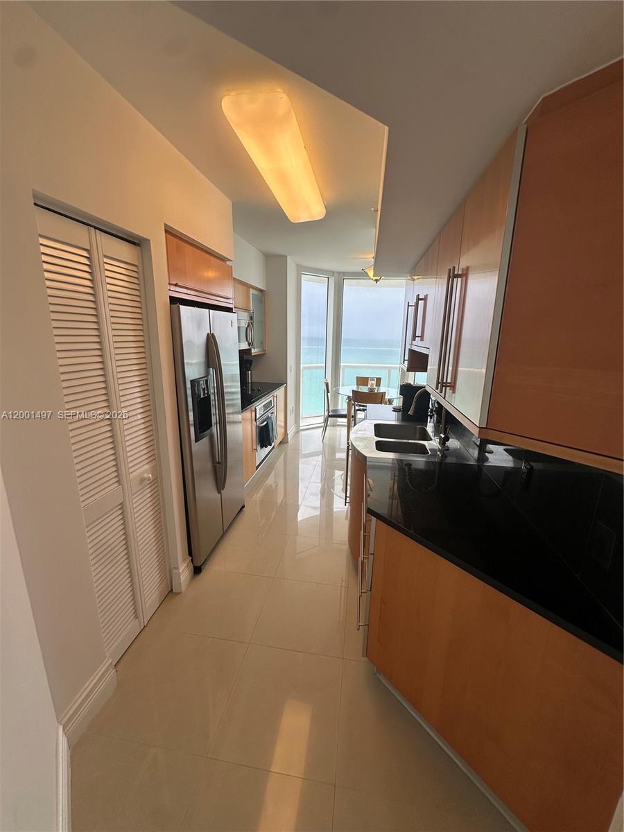 A galley kitchen features wood cabinetry, black countertops, stainless steel appliances, and a balcony view — 6365 Collins Ave # 1703, Miami Beach FL 33141, MIAMI BEACH, FL