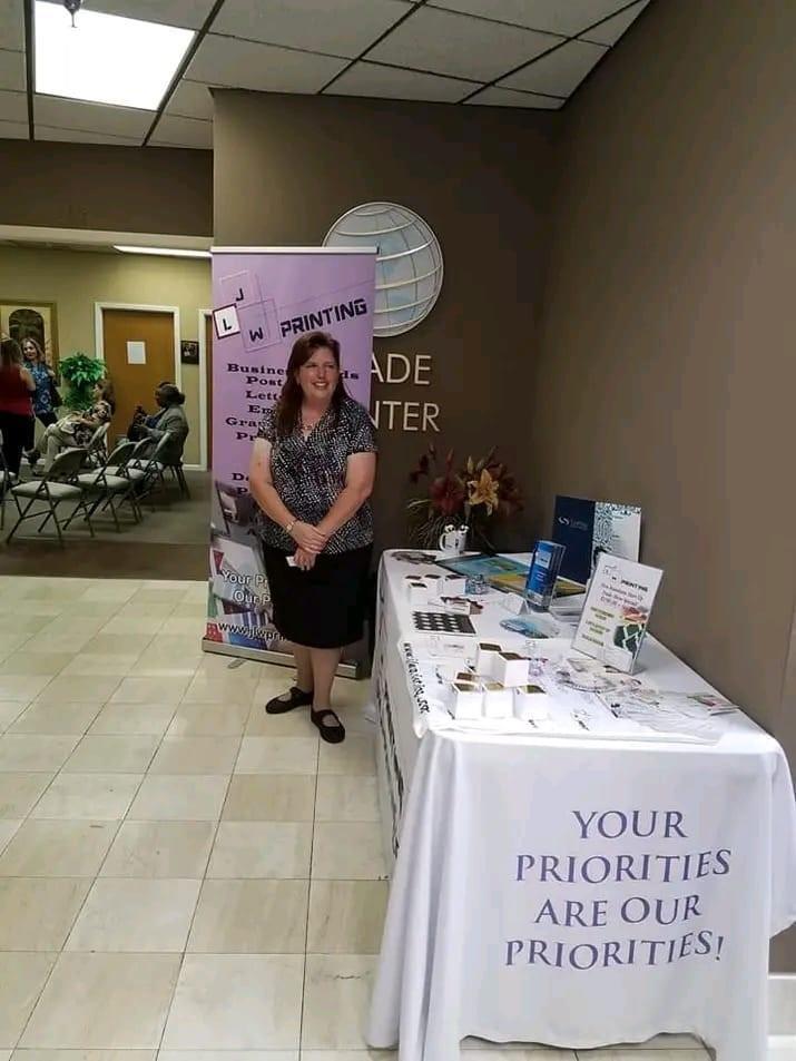 A woman stands behind a branded table at a business expo in an office building — 10001 Oakland Park Boulevard # 302, Sunrise FL 33351, SUNRISE, FL