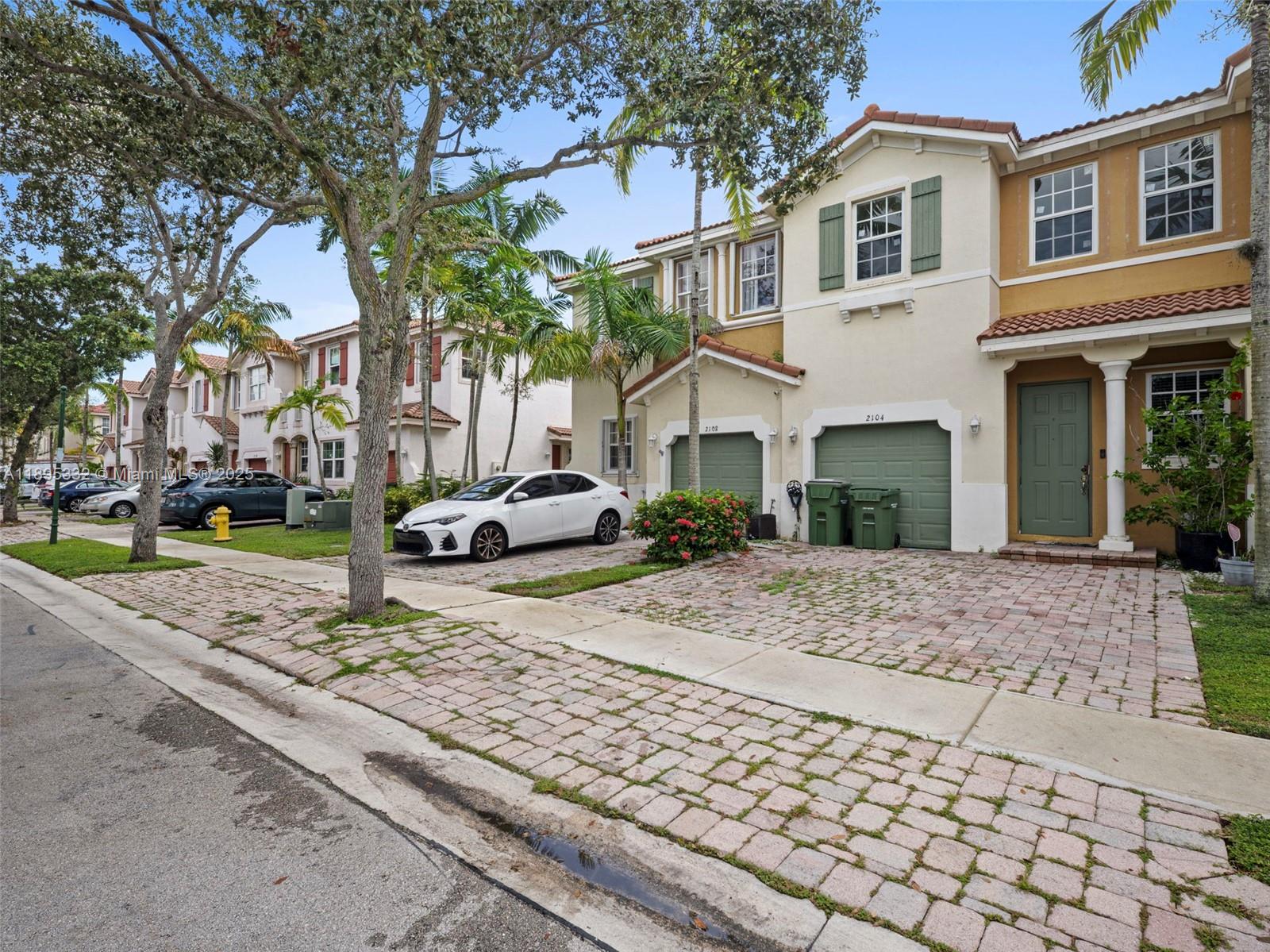 A street view of two-story townhomes with brick driveways, green garage doors, and parked cars — 2104 NE 3rd Ct, Homestead FL 33033, HOMESTEAD, FL