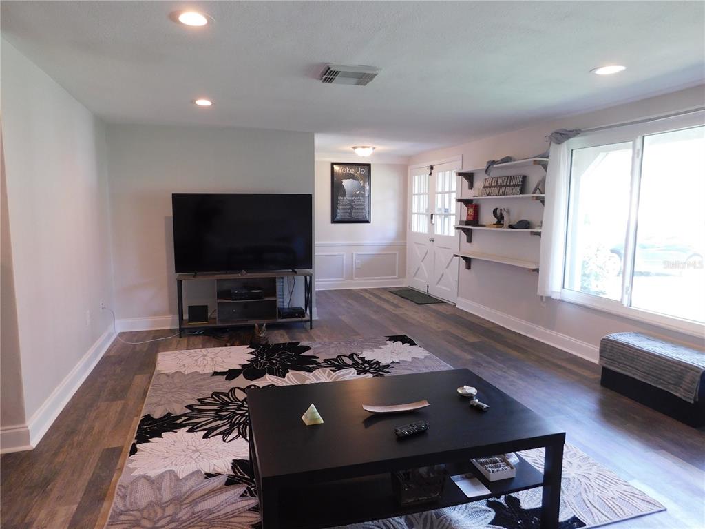 A living room features wood-look flooring, a large area rug, a television, and open shelving — 8020 HUNTERS WHIP ROW, HUDSON, FL
