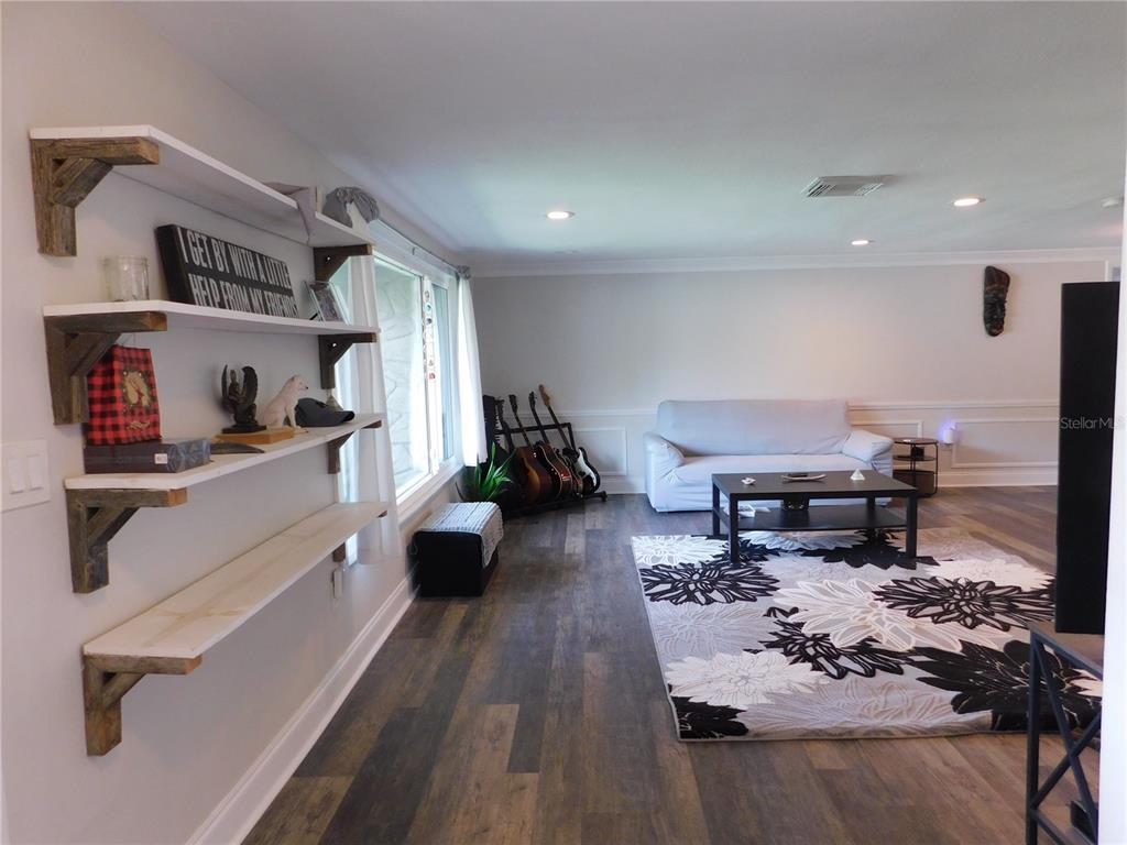 A living room features wood-look flooring, wall-mounted shelves, a sofa, and a floral area rug — 8020 HUNTERS WHIP ROW, HUDSON, FL