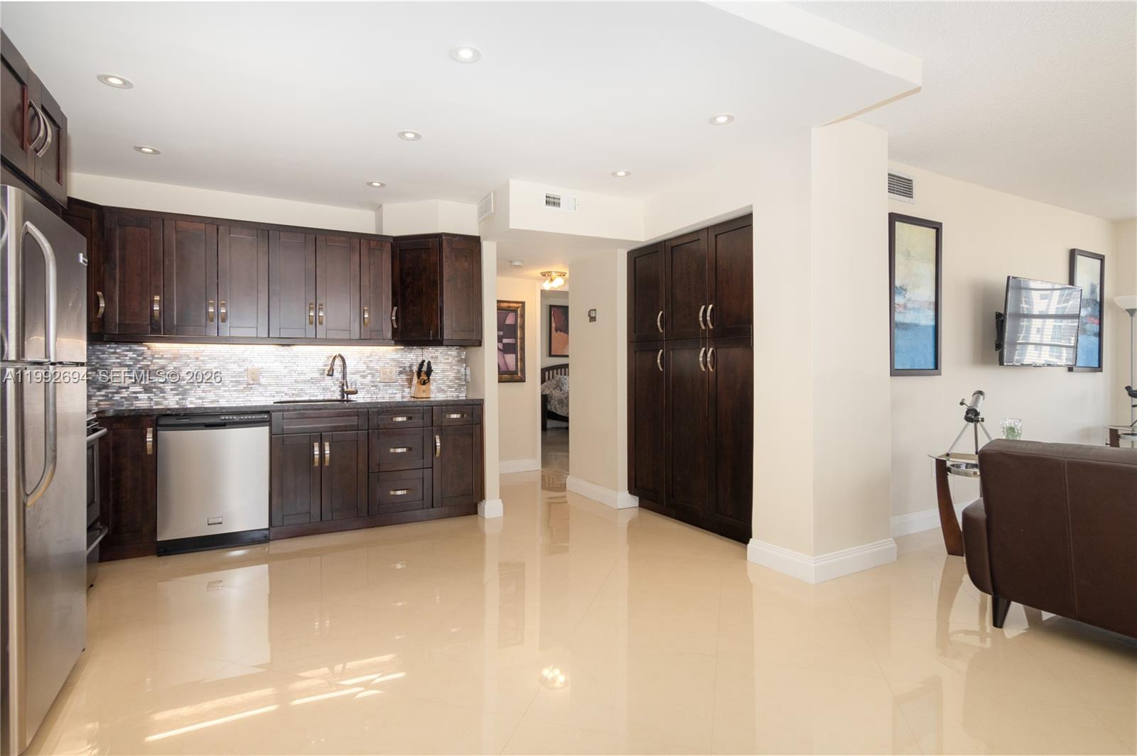 A kitchen with dark wood cabinets, stainless steel appliances, and tile flooring opens to a living area — 1965 S Ocean Dr # 9F, Hallandale Beach FL 33009, HALLANDALE BEACH, FL
