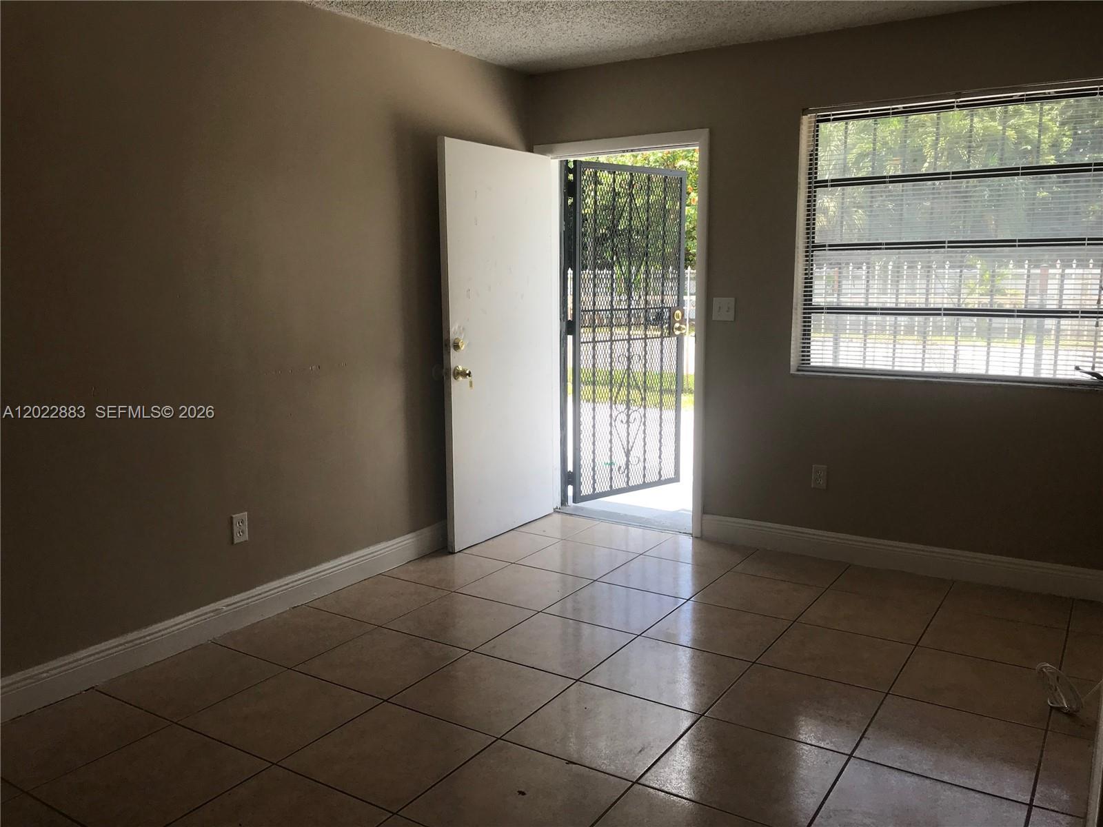 An empty room features beige walls, tiled flooring, a white door, and a metal security gate — 13315 Alexandria Dr # 3, Opa-Locka FL 33054, OPA-LOCKA, FL