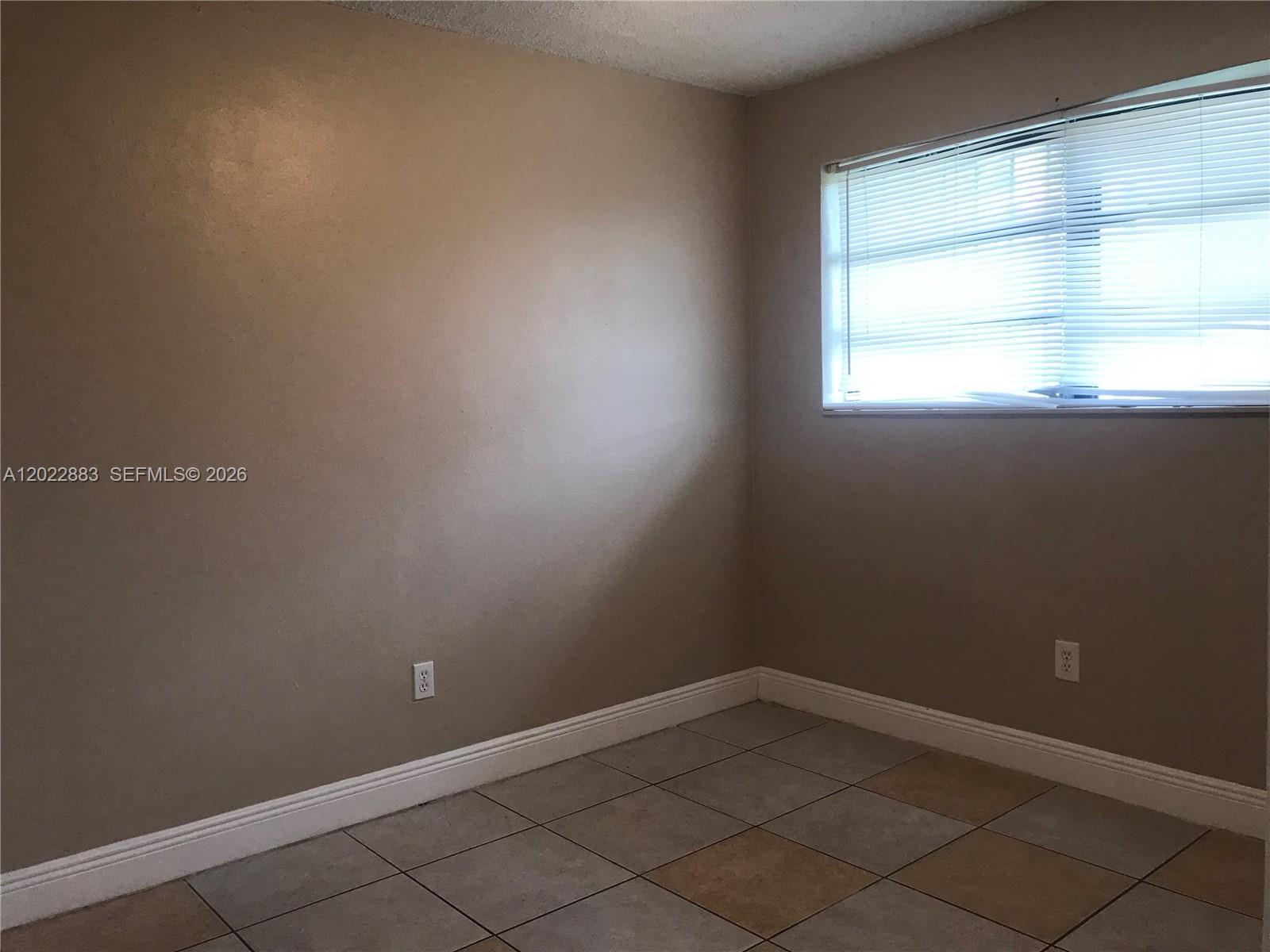 An empty room features beige walls, white baseboards, tiled flooring, and a window with blinds — 13315 Alexandria Dr # 3, Opa-Locka FL 33054, OPA-LOCKA, FL