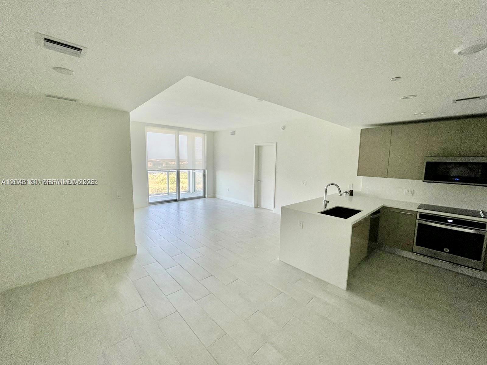 An open-concept kitchen with modern cabinetry and white tile floors overlooks a bright living area — 2000 Metropica Way # 1602, Sunrise FL 33323, SUNRISE, FL
