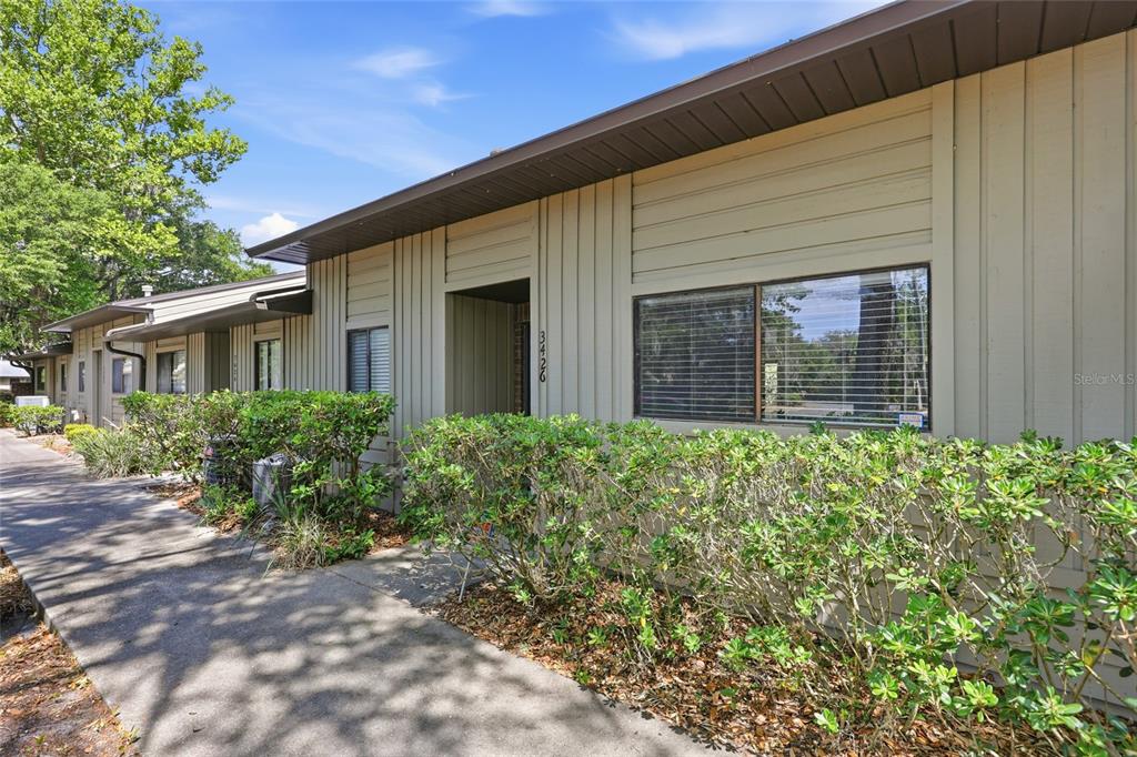 A tan, single-story exterior with vertical siding, a concrete walkway, and a landscaped hedge — 3426 NW 37TH AVENUE UNIT 3426, GAINESVILLE, FL
