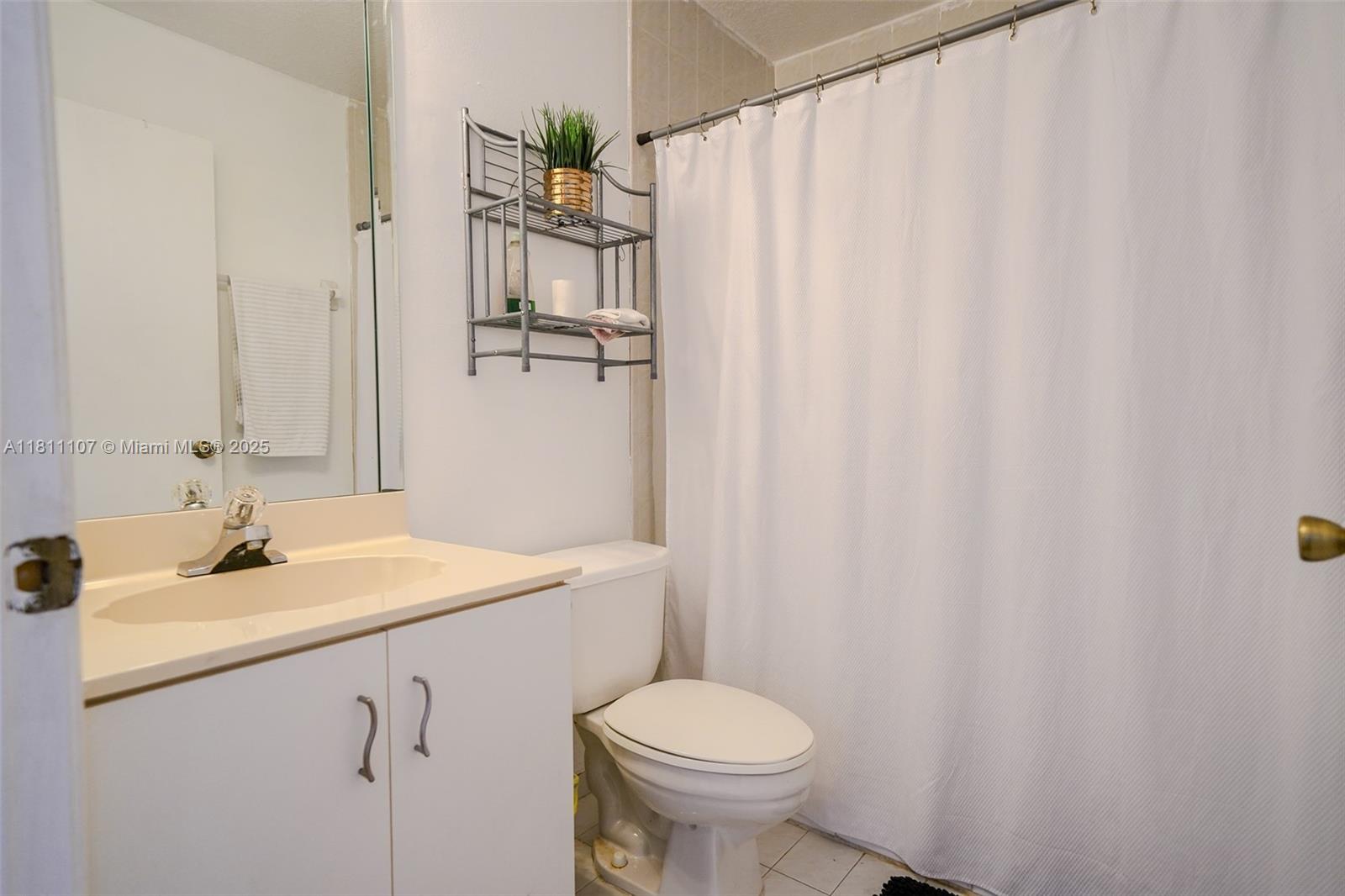 A bathroom features a white vanity, a toilet, a wall-mounted metal shelf, and a shower curtain — 14816 SW 104th St # 85, Miami FL 33196, MIAMI, FL