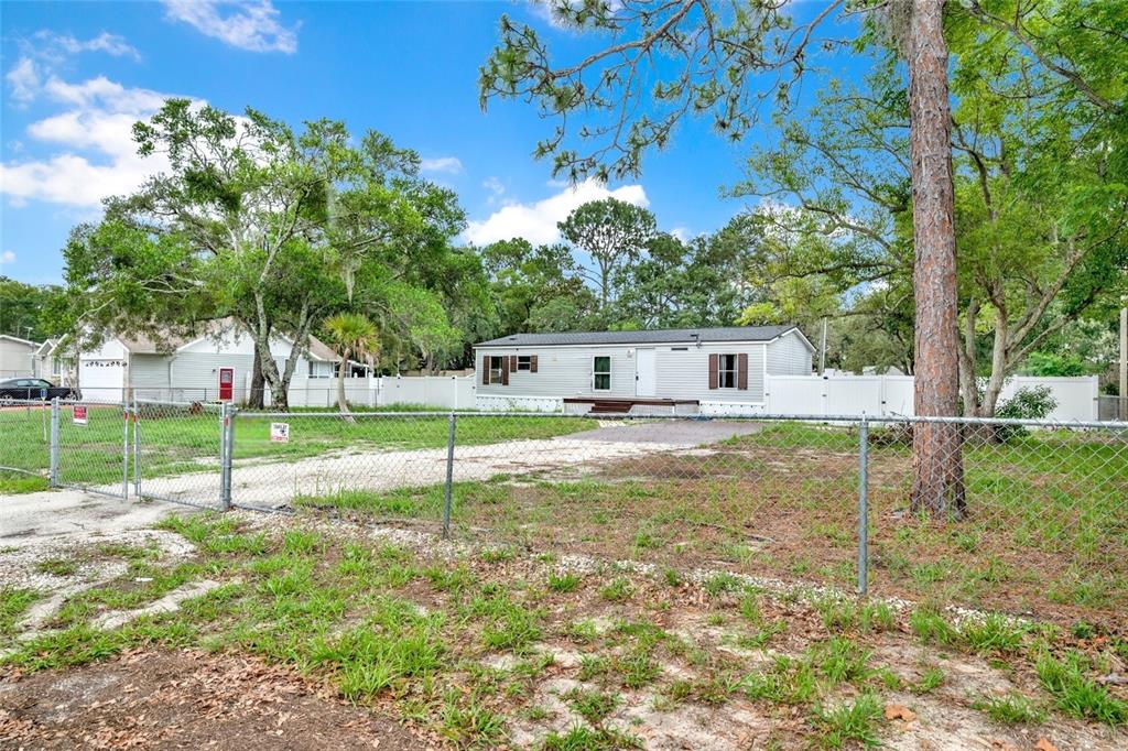 A single-story manufactured home sits on a fenced lot with trees under a blue sky — 7194 SEALAWN DRIVE, SPRING HILL, FL