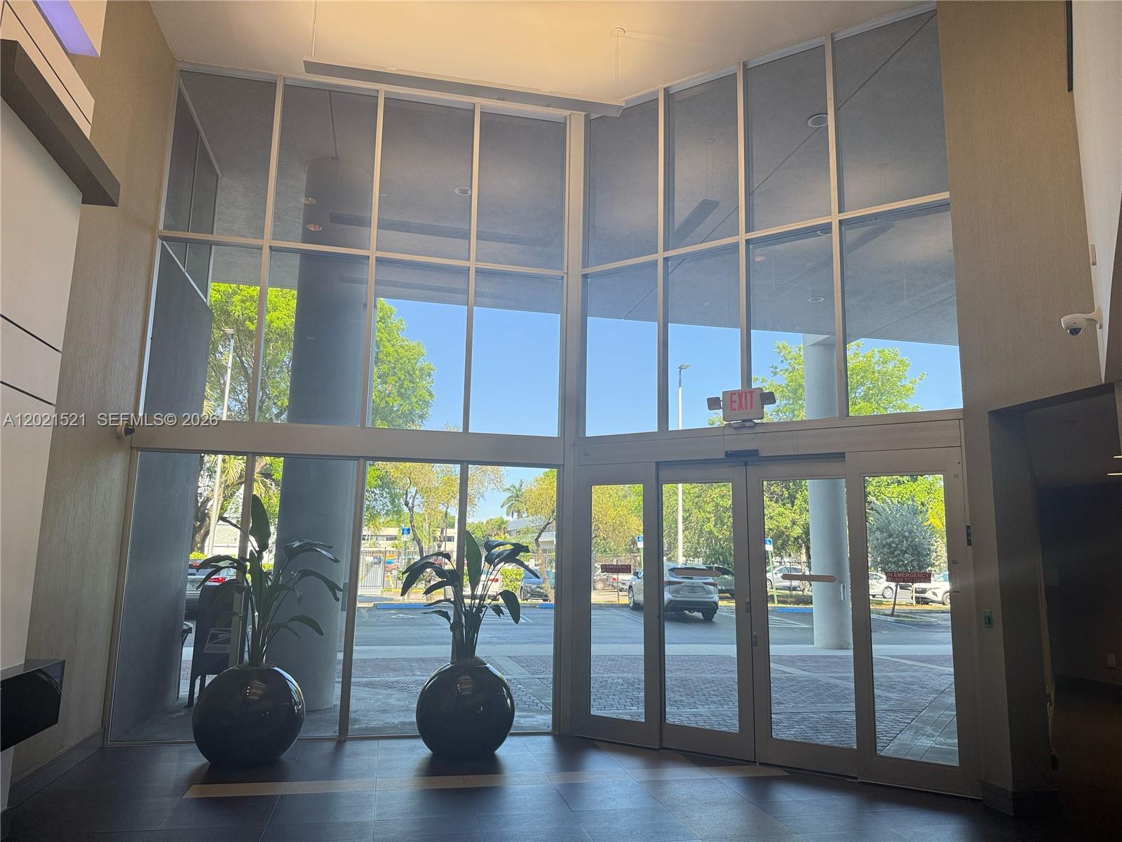 A modern building lobby features floor-to-ceiling windows, glass entry doors, and two large potted plants — 290 NW 165th St # 5C, Miami FL 33169, MIAMI, FL