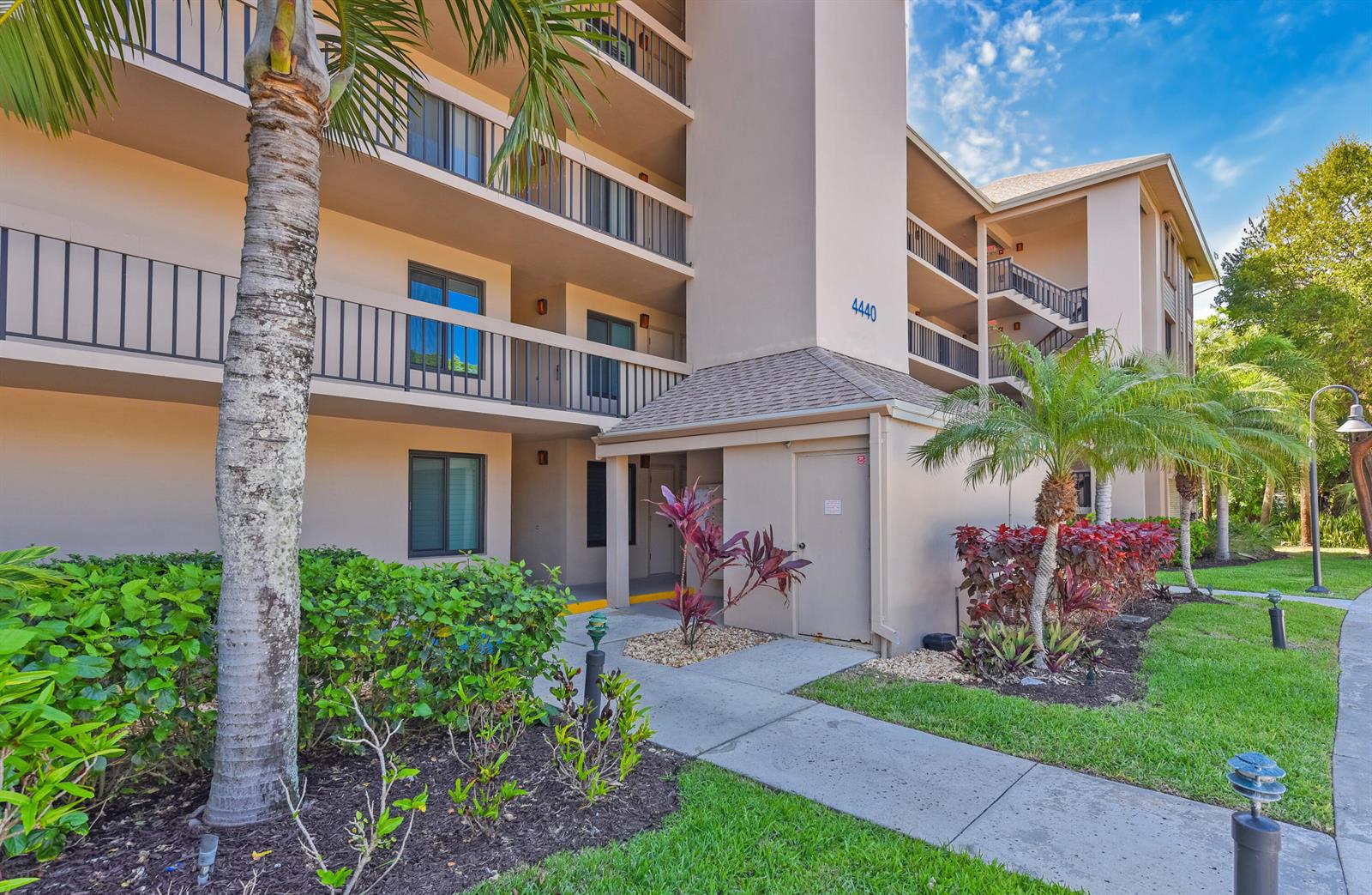 Exterior view of a multi-story apartment building with balconies, palm trees, and a concrete walkway — 4440 NE Sandpebble Trace # 104, Stuart FL 34996, STUART, FL