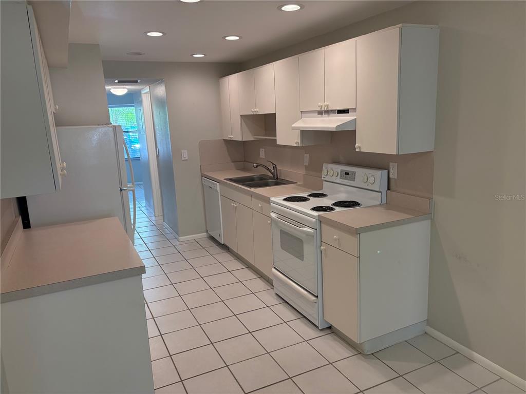 A galley-style kitchen features white cabinets, light countertops, white appliances, and tiled flooring — 515 30TH AVENUE W UNIT H108, BRADENTON, FL