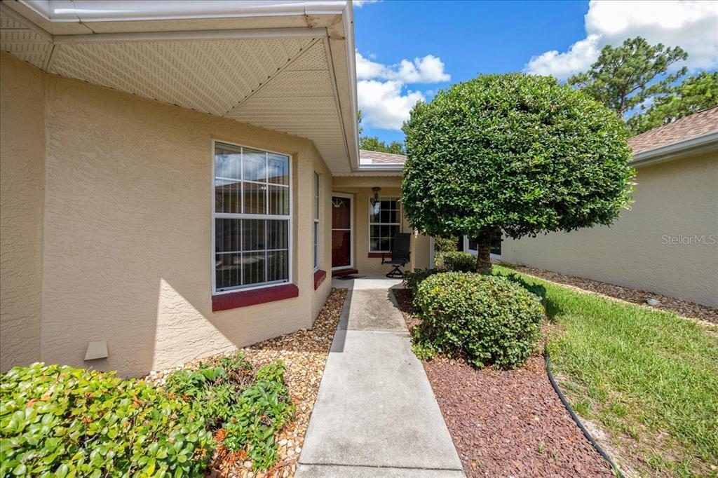 A concrete walkway leads to the front entrance of a beige house with landscaping — 9082 SW 102ND CIRCLE, OCALA, FL