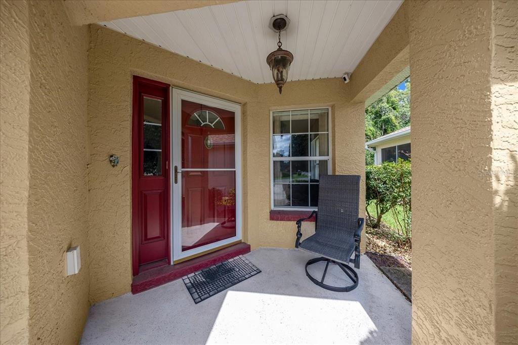 A covered front porch features a red door with a storm door and a chair — 9082 SW 102ND CIRCLE, OCALA, FL
