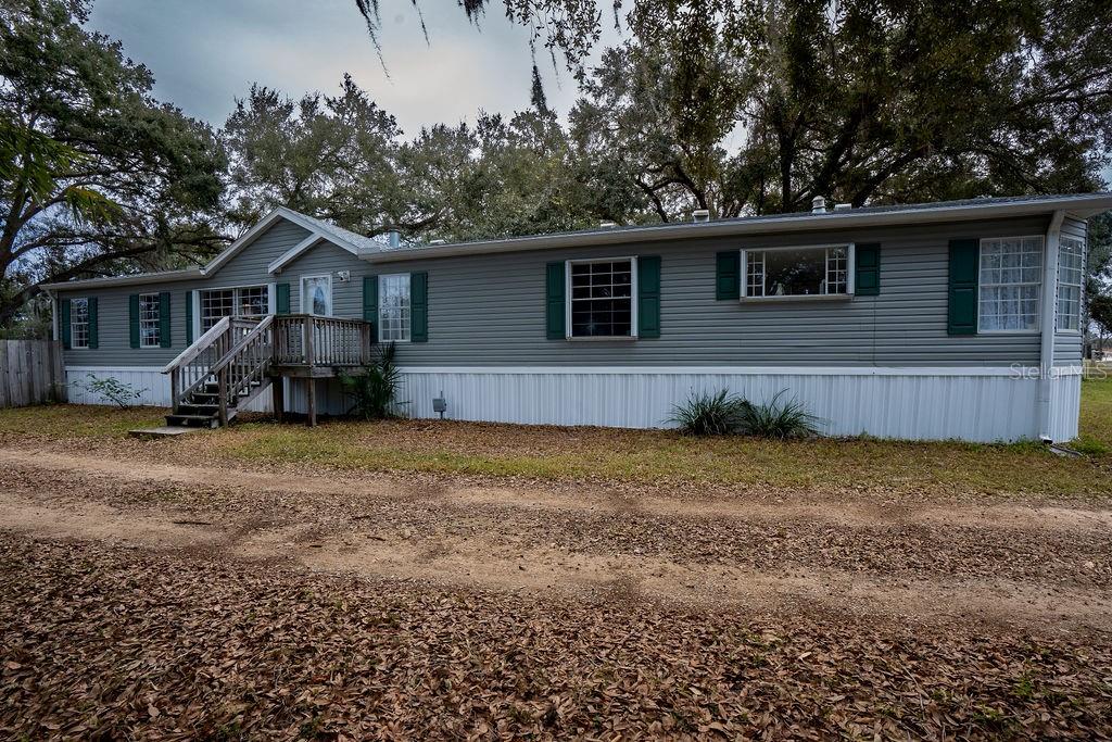 A gray manufactured home with green shutters and a wooden deck sits on a dirt lot — 13811 COUNTY ROAD 109D, LADY LAKE, FL