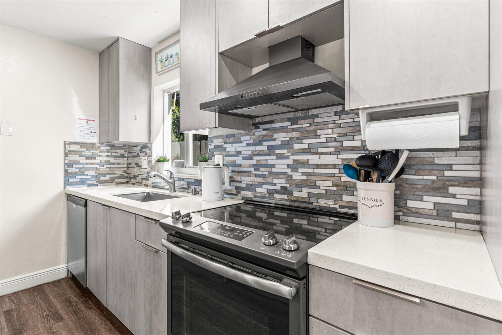 A modern kitchen features gray cabinets, a glass-top stove, and a blue-toned mosaic tile backsplash — 1121 N 66th Terrace, Hollywood FL 33024, HOLLYWOOD, FL