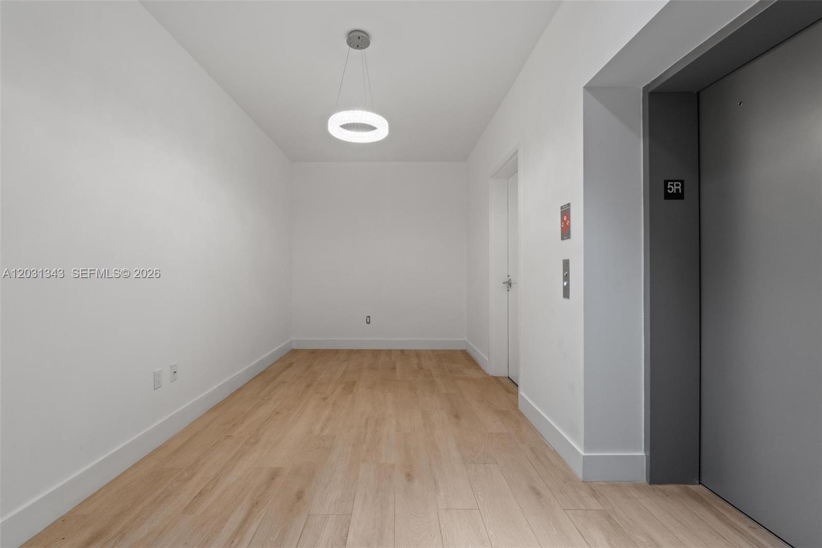An empty room features white walls, light wood flooring, a circular pendant light, and an elevator — NORTH MIAMI BEACH, FL