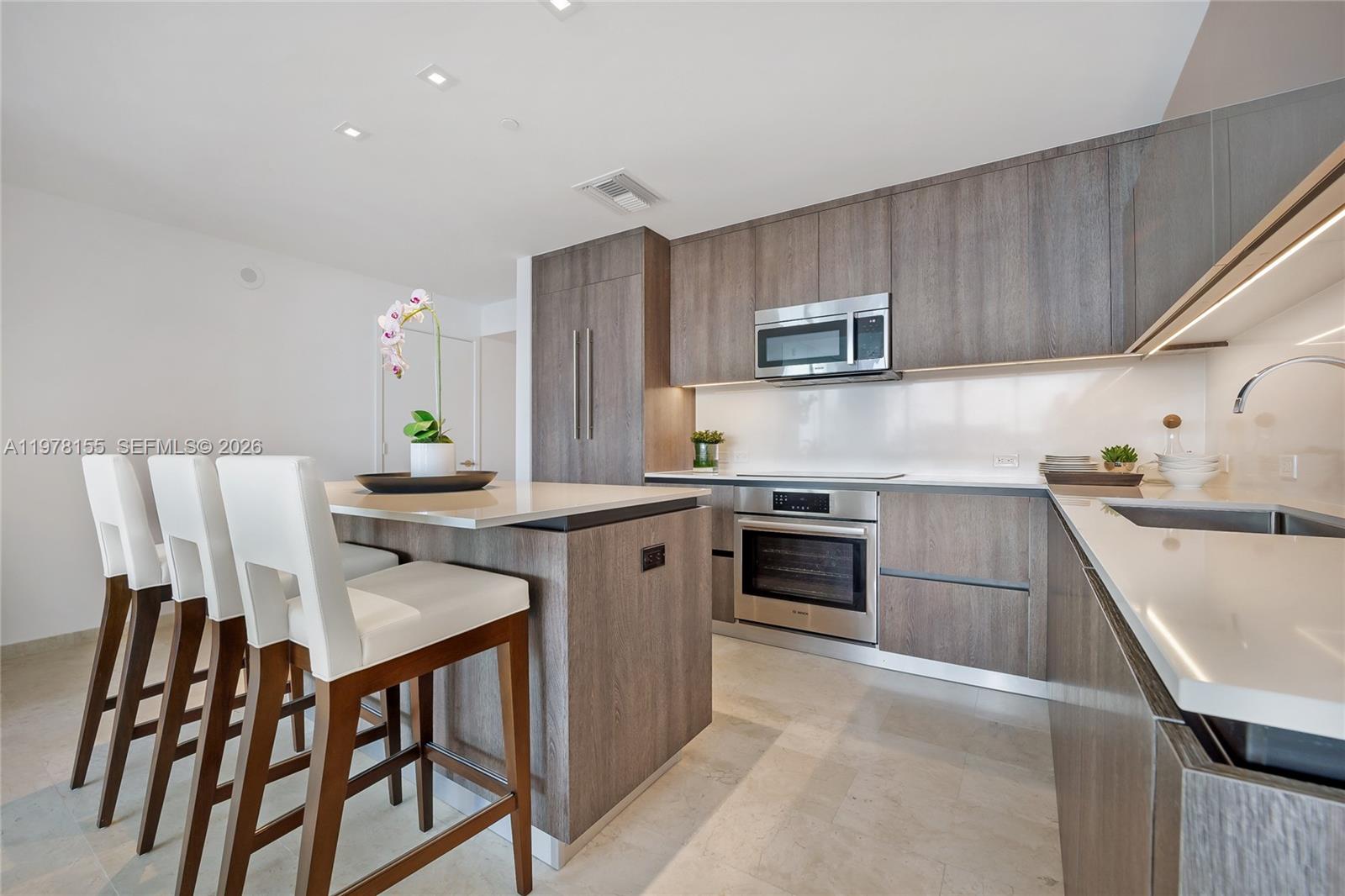 A modern kitchen features wood-grain cabinetry, white countertops, a central island, and three bar stools — 88 SW 7th St # 1810, Miami FL 33130, MIAMI, FL