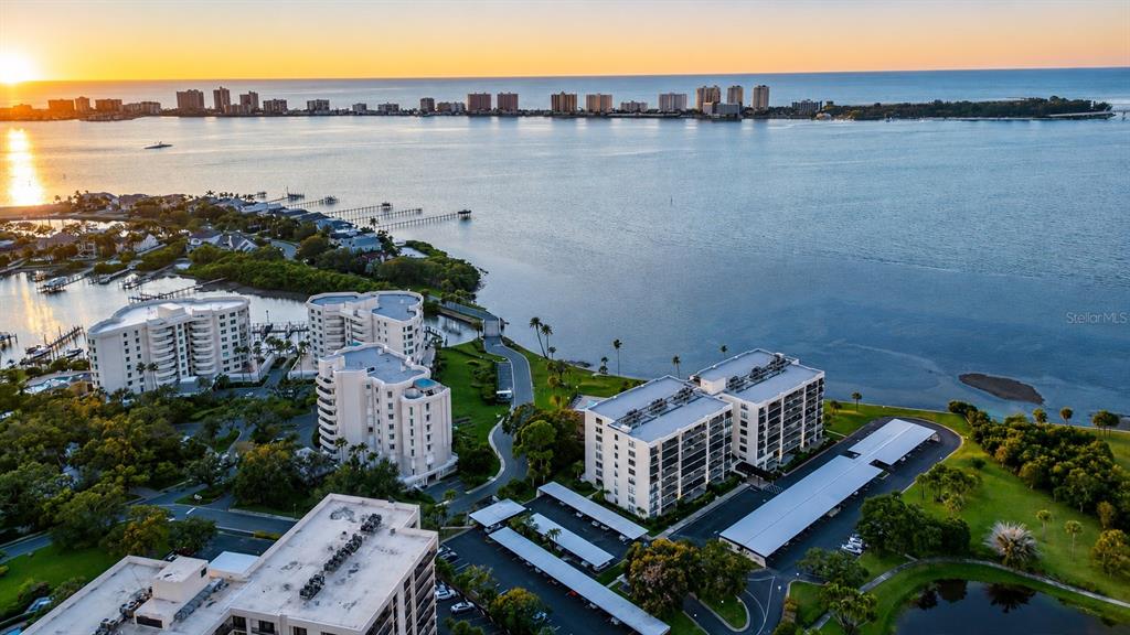 An aerial view shows waterfront apartment buildings, parking structures, and a sunset over the ocean — 100 OAKMONT LANE UNIT 507, BELLEAIR, FL
