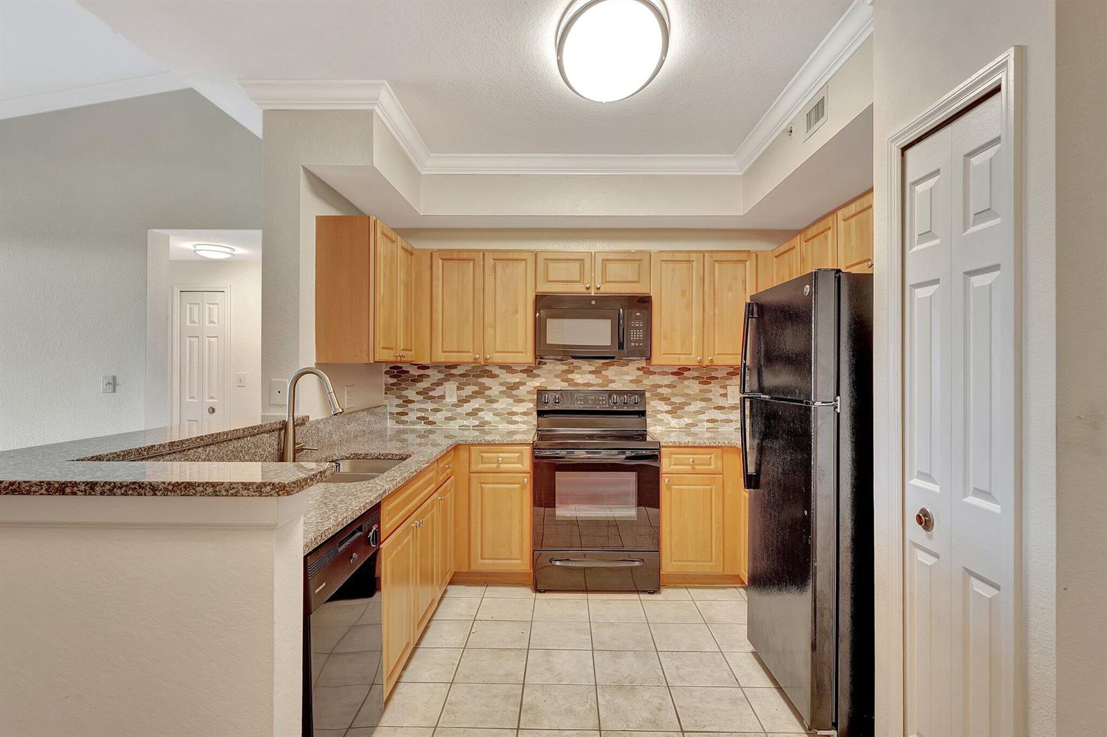 A kitchen features light wood cabinets, black appliances, granite countertops, and a tiled floor — 15055 Michelangelo Boulevard # 208, Delray Beach FL 33446, DELRAY BEACH, FL