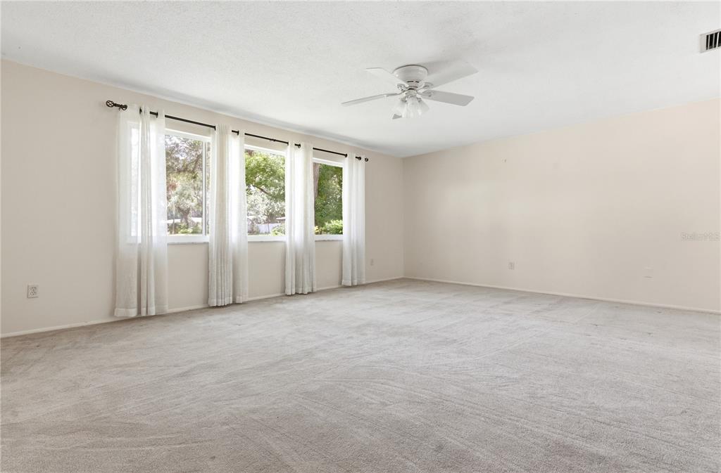 A spacious, carpeted living room features a ceiling fan and three windows with white curtains — 1904 GRANADA COURT, CLEARWATER, FL