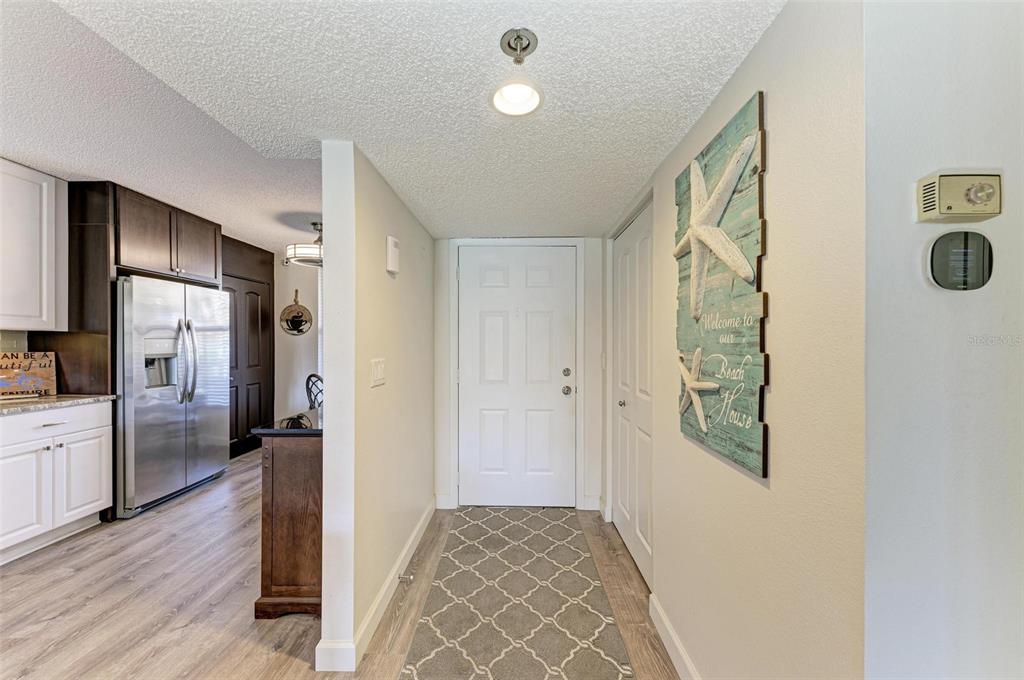 A hallway with wood-look flooring leads to a white front door near a kitchen — 4480 FAIRWAYS BOULEVARD UNIT 109, BRADENTON, FL