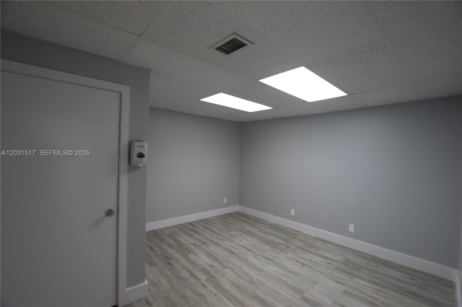 An empty room features gray walls, wood-look flooring, a drop ceiling, and a white door — HOLLYWOOD, FL