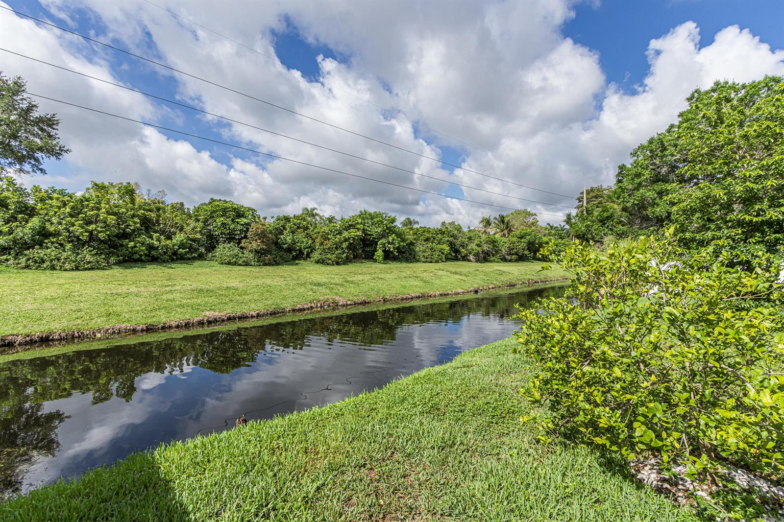 A narrow canal flows between grassy banks lined with trees under a cloudy blue sky — 16942 SW 5th Way, Weston FL 33326, WESTON, FL