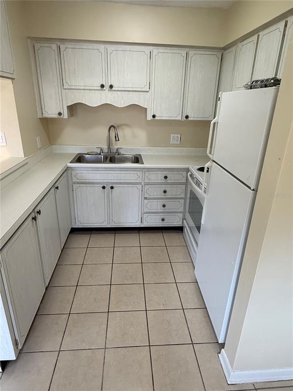 A small kitchen features white cabinets, a double-basin sink, a white refrigerator, and tiled flooring — 2099 KINGS HWY #1 UNIT 1, CLEARWATER, FL