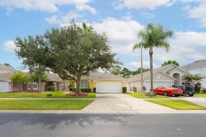 A yellow house with a two-car garage and a large tree in the front yard — 1017 LAKE BERKLEY DR, KISSIMMEE, FL
