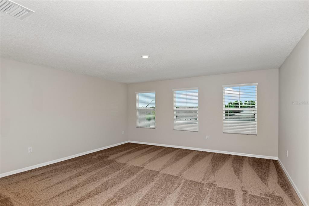 An empty bedroom with beige walls, brown carpet, and three windows with white blinds — 31670 BARONNE STREET, WESLEY CHAPEL, FL