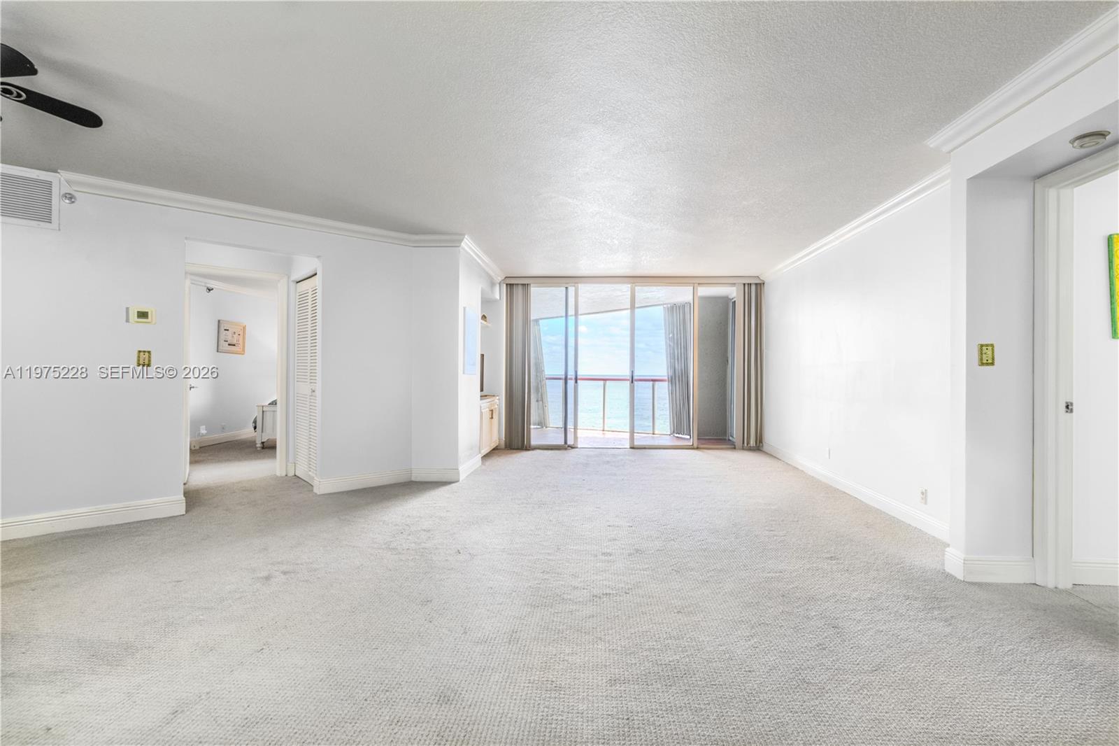 An empty living room with white walls, beige carpeting, and a balcony overlooking the ocean — 6767 Collins Ave # 706, Miami Beach FL 33141, MIAMI BEACH, FL