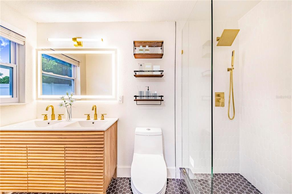 A modern bathroom features a double vanity, a glass-enclosed shower, and gold-toned fixtures — 3104 ASH COURT, DUNEDIN, FL