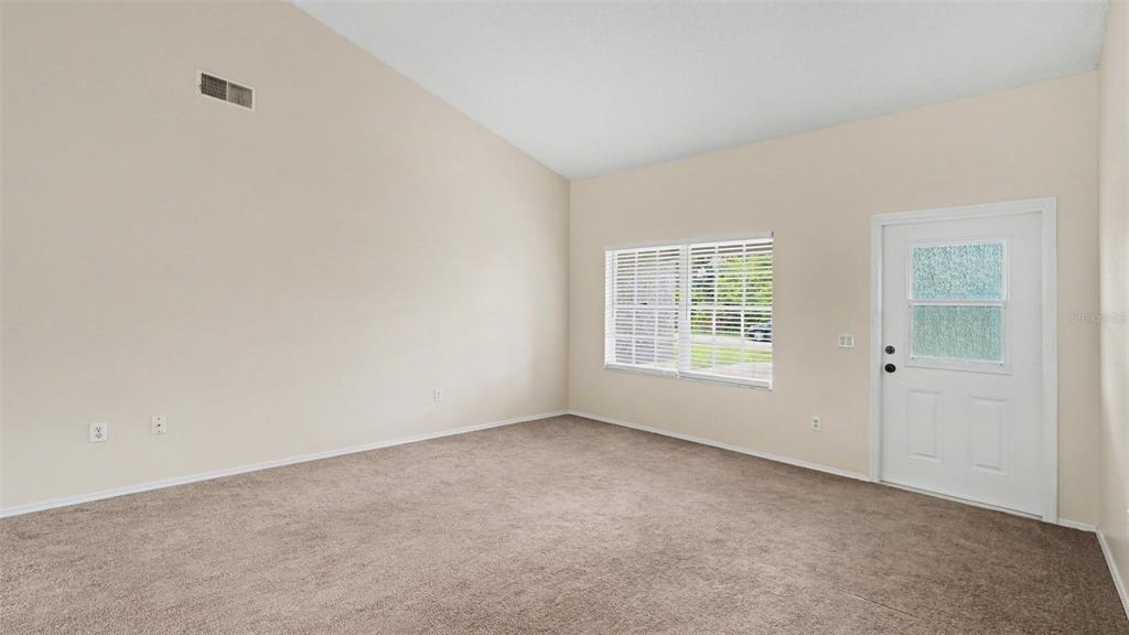 An empty living room with beige walls, tan carpeting, a vaulted ceiling, and a white door — 206 OAK CIRCLE, OCALA, FL