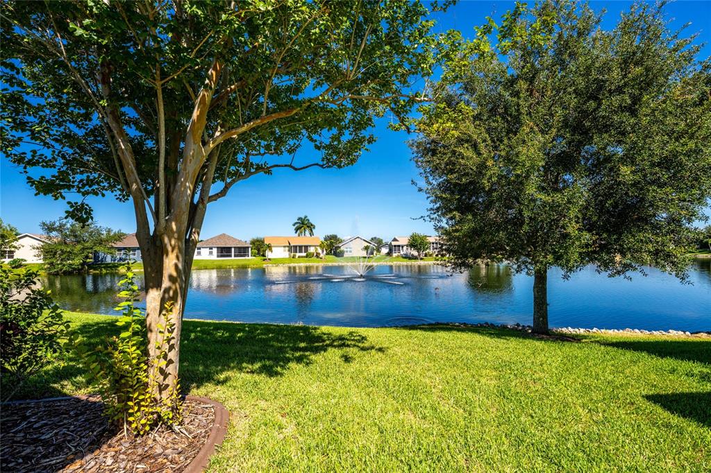 A grassy lawn leads to a pond with a fountain, framed by trees and houses — 24819 RIO VILLA LAKES CIRCLE, PUNTA GORDA, FL