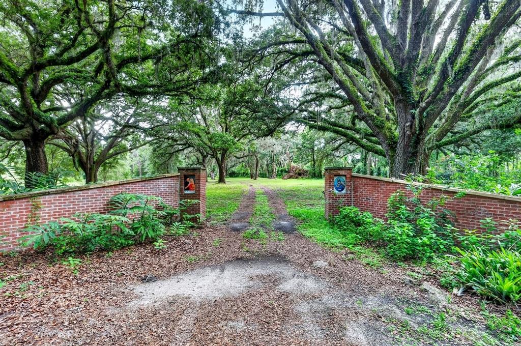 A brick-walled entrance leads to a dirt driveway shaded by large, moss-covered oak trees — 1980 LAKE HIRES ROAD, DE LEON SPRINGS, FL