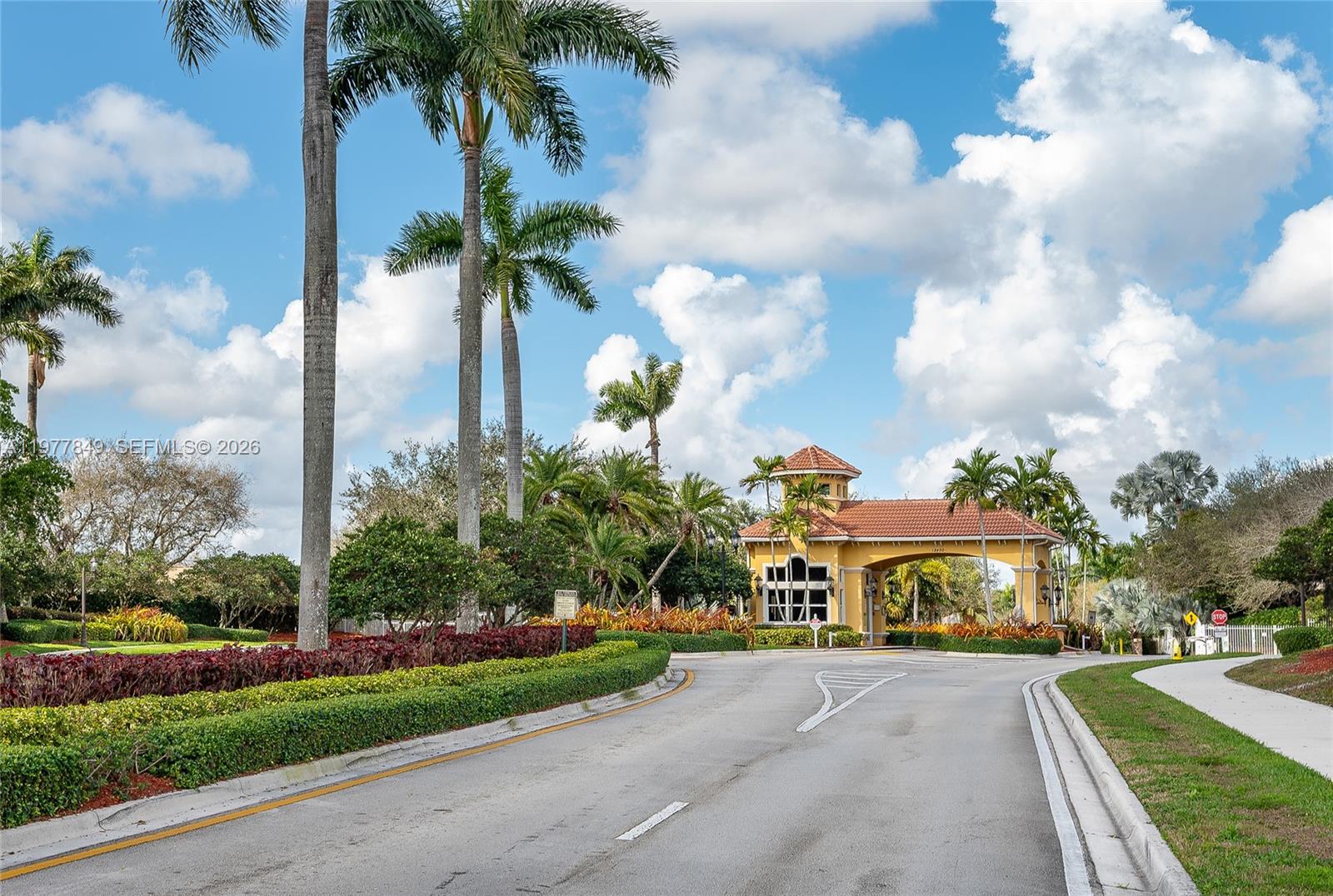 A paved road leads toward a gated community entrance with a yellow guardhouse and palm trees — 13391 SW 42nd St, Miramar FL 33027, MIRAMAR, FL