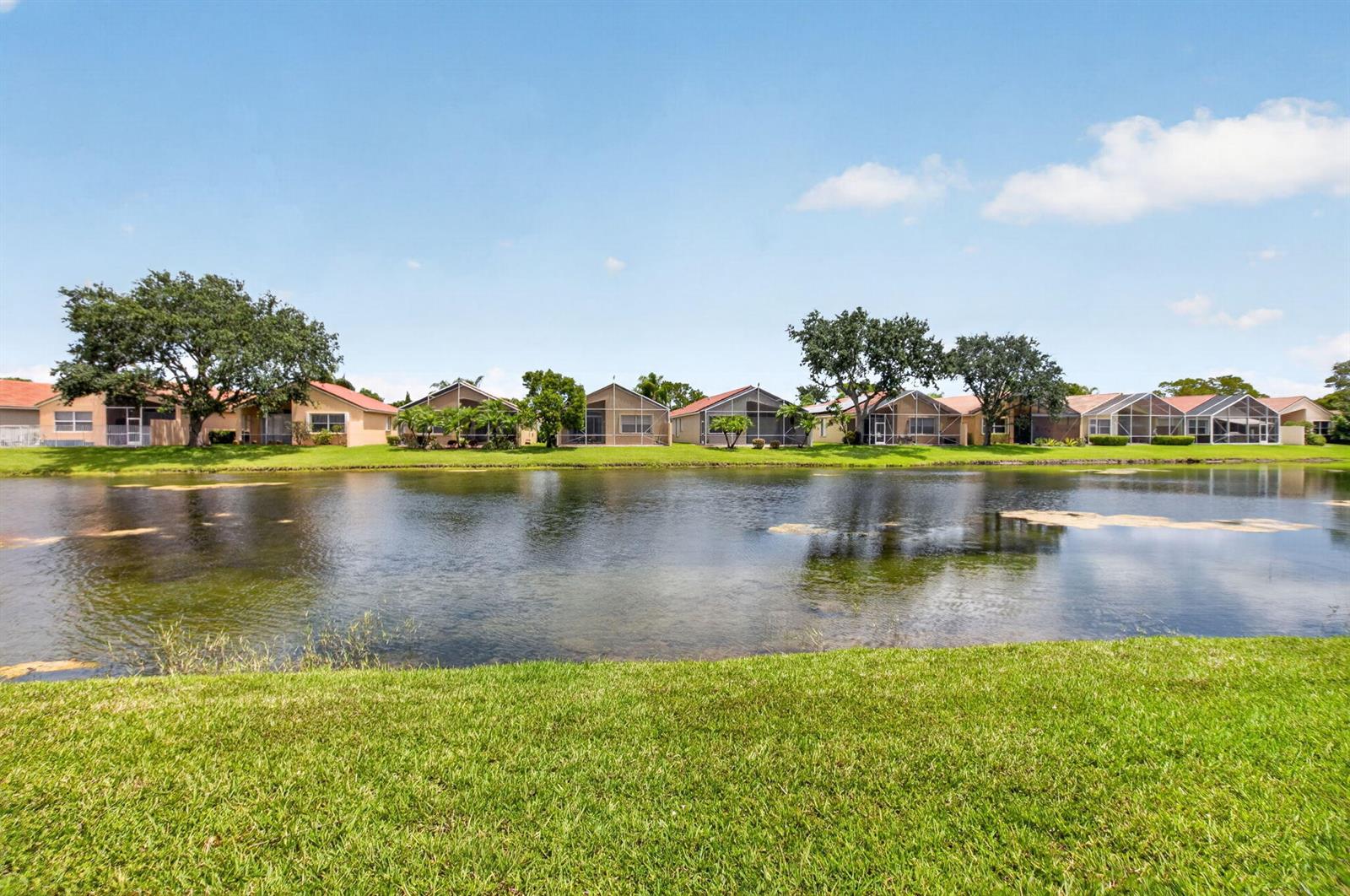 A row of houses with screened lanais sits across a pond behind a grassy lawn — 7035 Vivaldi Lane, Delray Beach FL 33446, DELRAY BEACH, FL